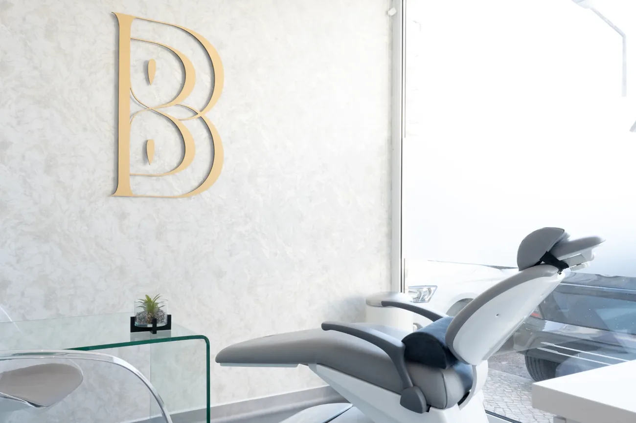 Espaço Beleza Infinita: parede de estuque claro com o monograma dourado da marca, cadeira de tratamento e mesa de vidro junto a uma janela ampla